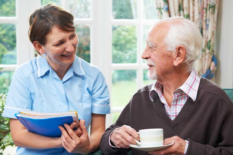 Two senior adults sit at a dining table with a caregiver, sharing a meal and conversation in a bright, home-like room.